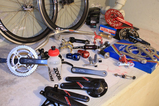 A collection of used bicycle components and parts laid out on a light-colored workbench, including a white ZOOM CRANER crank arm, black carbon fiber pedals with red branding, a red chainring, and other bicycle hardware.